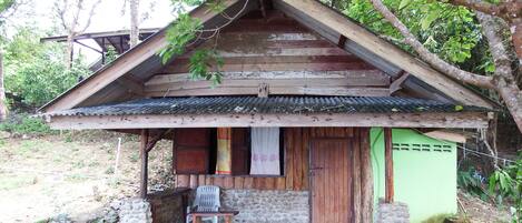 A rustic wooden cabin with a sloped roof, a porch with a single chair, and a stone wall foundation.