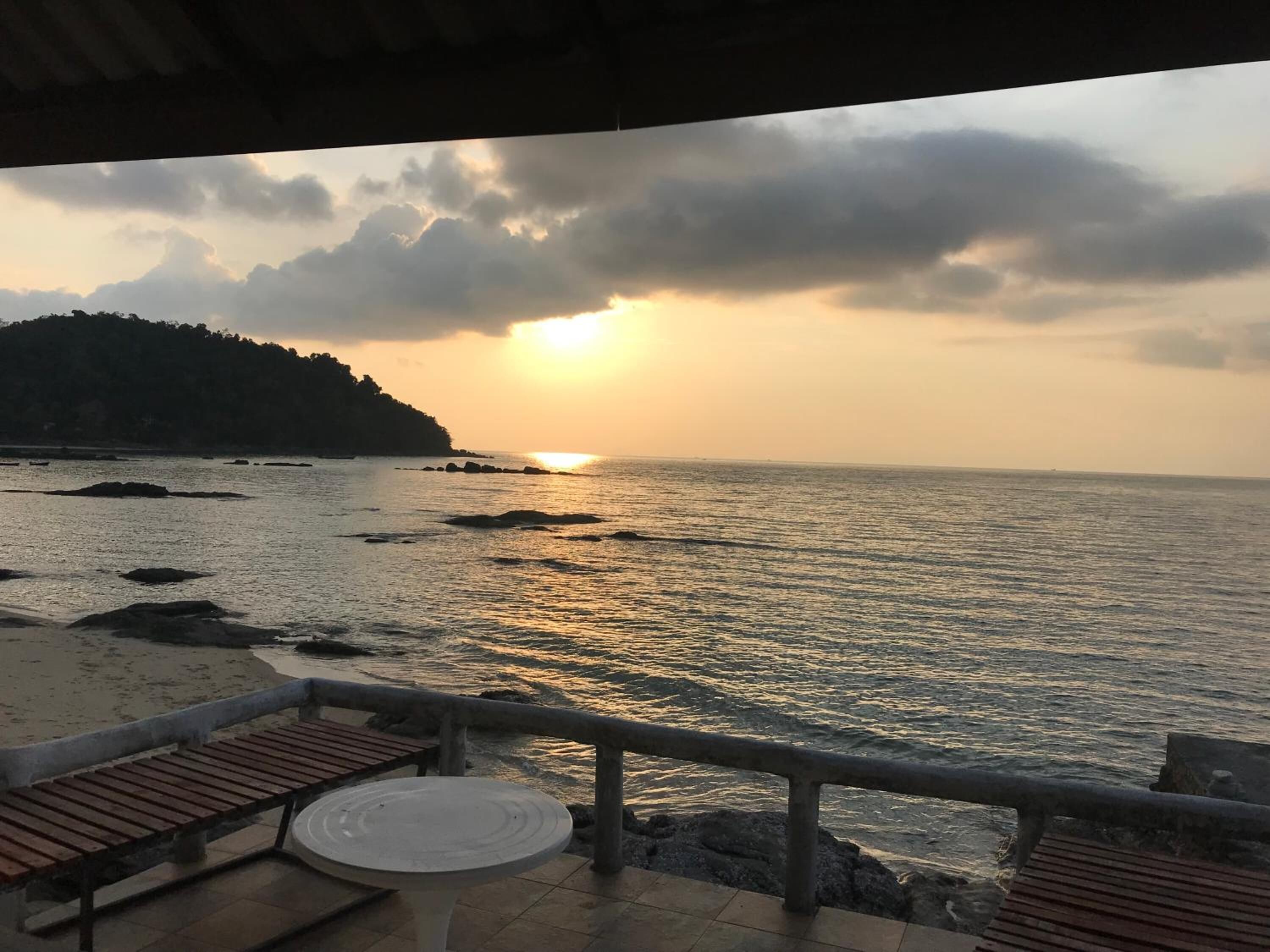 A seaside view with a sunset, rocky shoreline, and a small table and bench on a terrace.