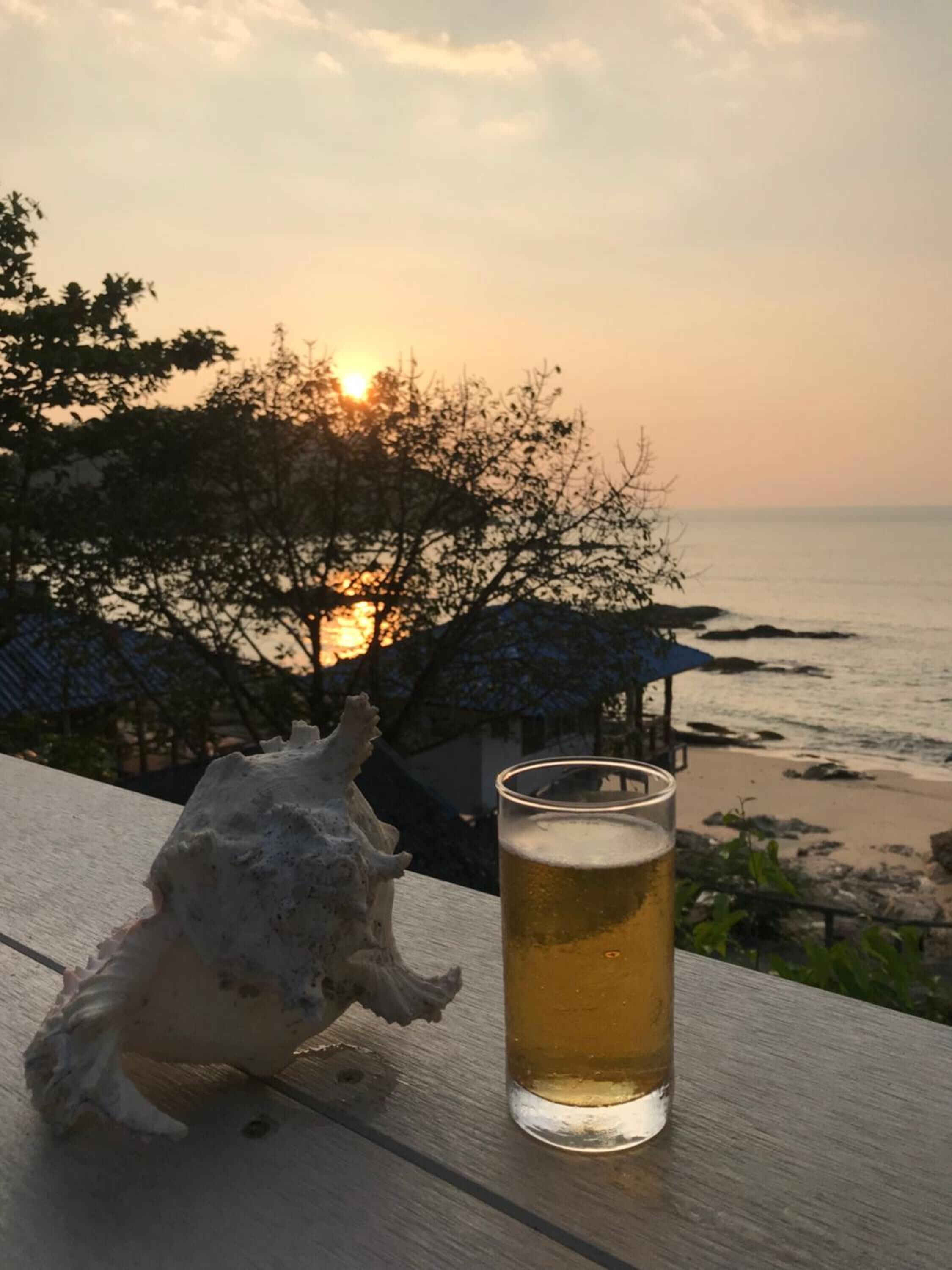A glass of beer on a wooden table with a beach and sunset in the background.