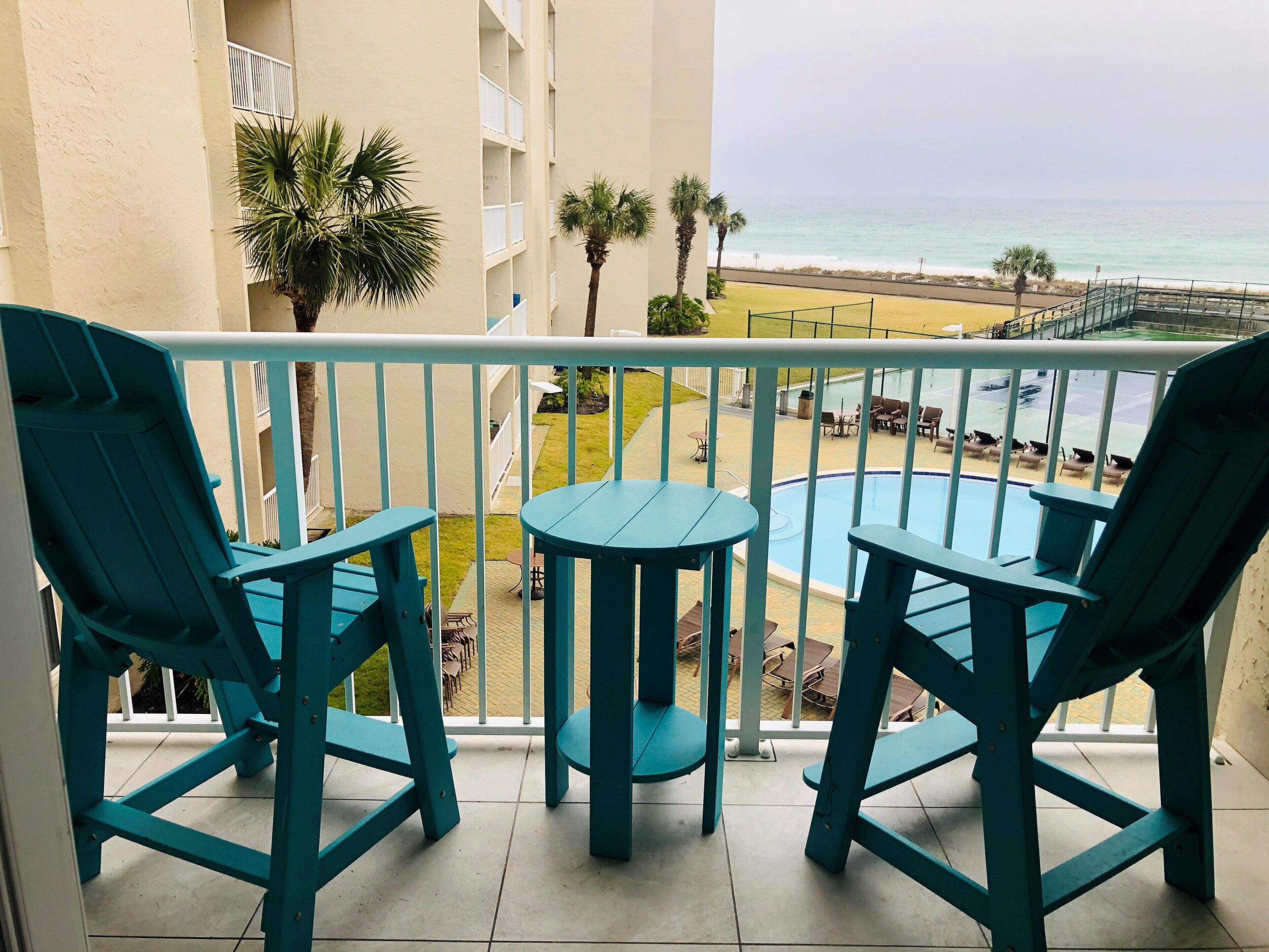 A balcony with teal chairs and a small table, overlooking a pool and beach area.