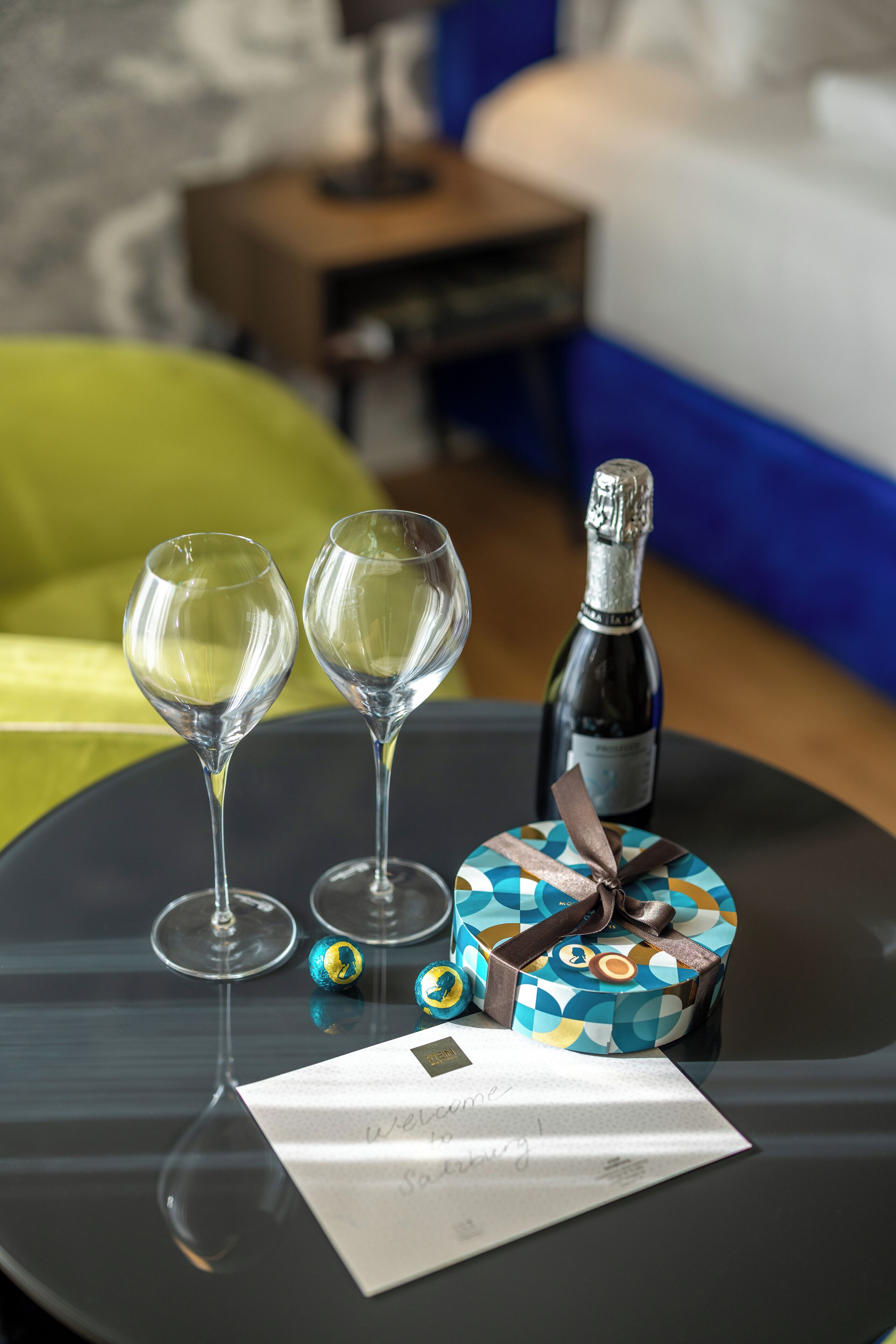 Una mesa con dos copas de vino, una botella de champán, una caja de regalo y una nota sobre una superficie reflectante.