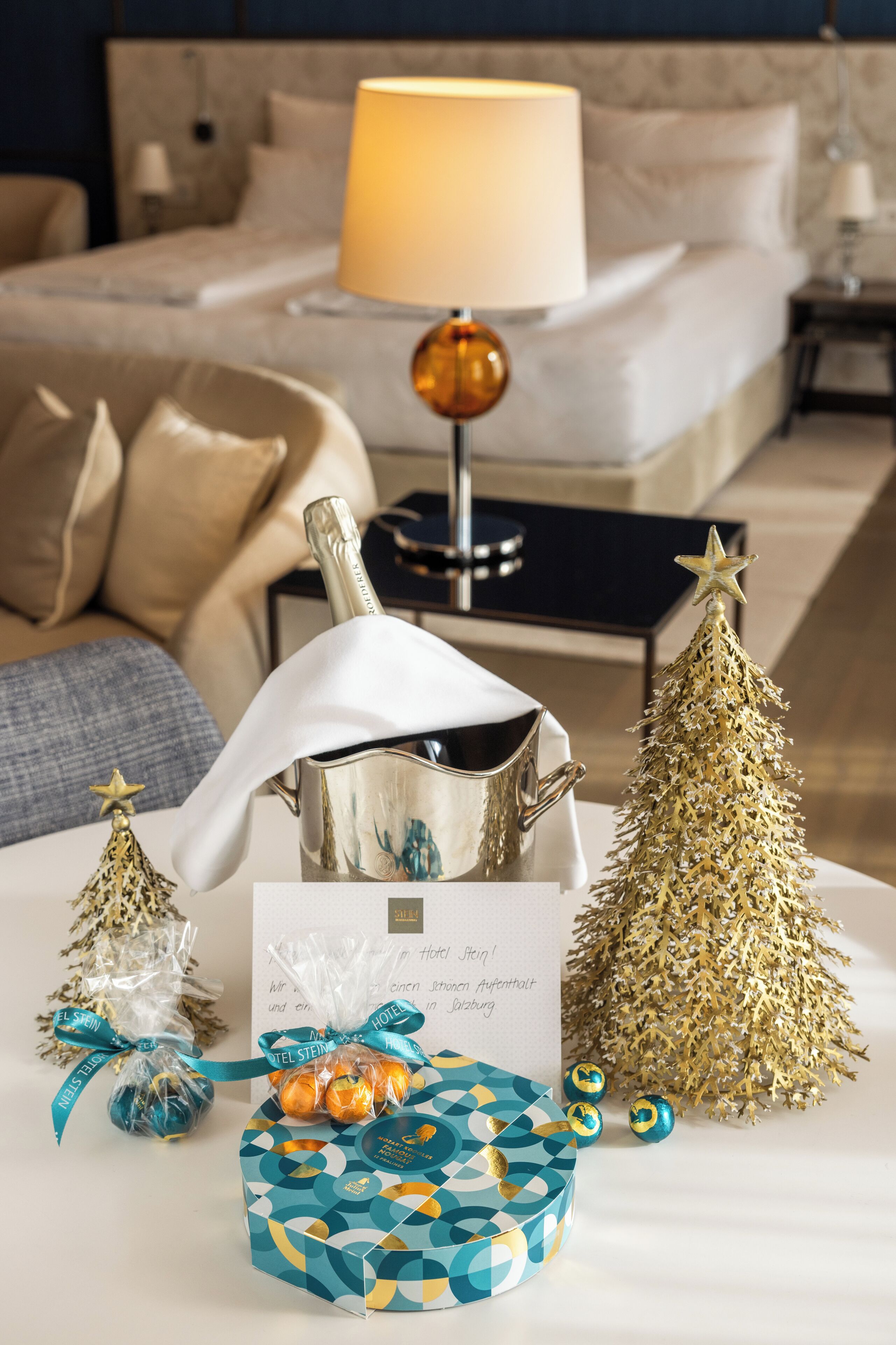 A hotel room with a bed, a nightstand, a lamp, a decorative bottle, a Christmas tree, and a gift box.