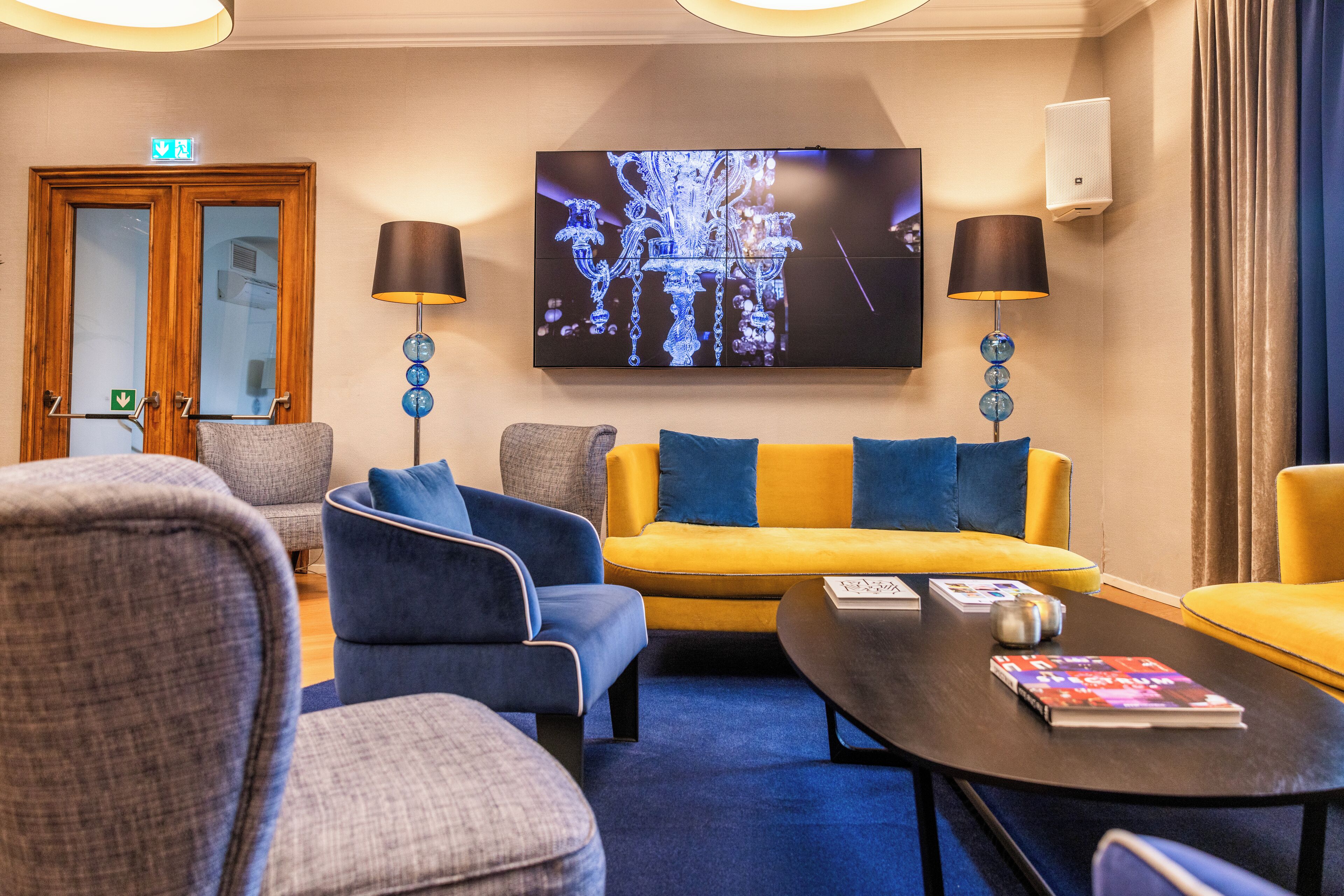 Un hall de hotel con diseño moderno, que presenta un televisor de pantalla plana grande montado en la pared, un sofá con cojines azules y amarillos, y dos lámparas con bases azules.
