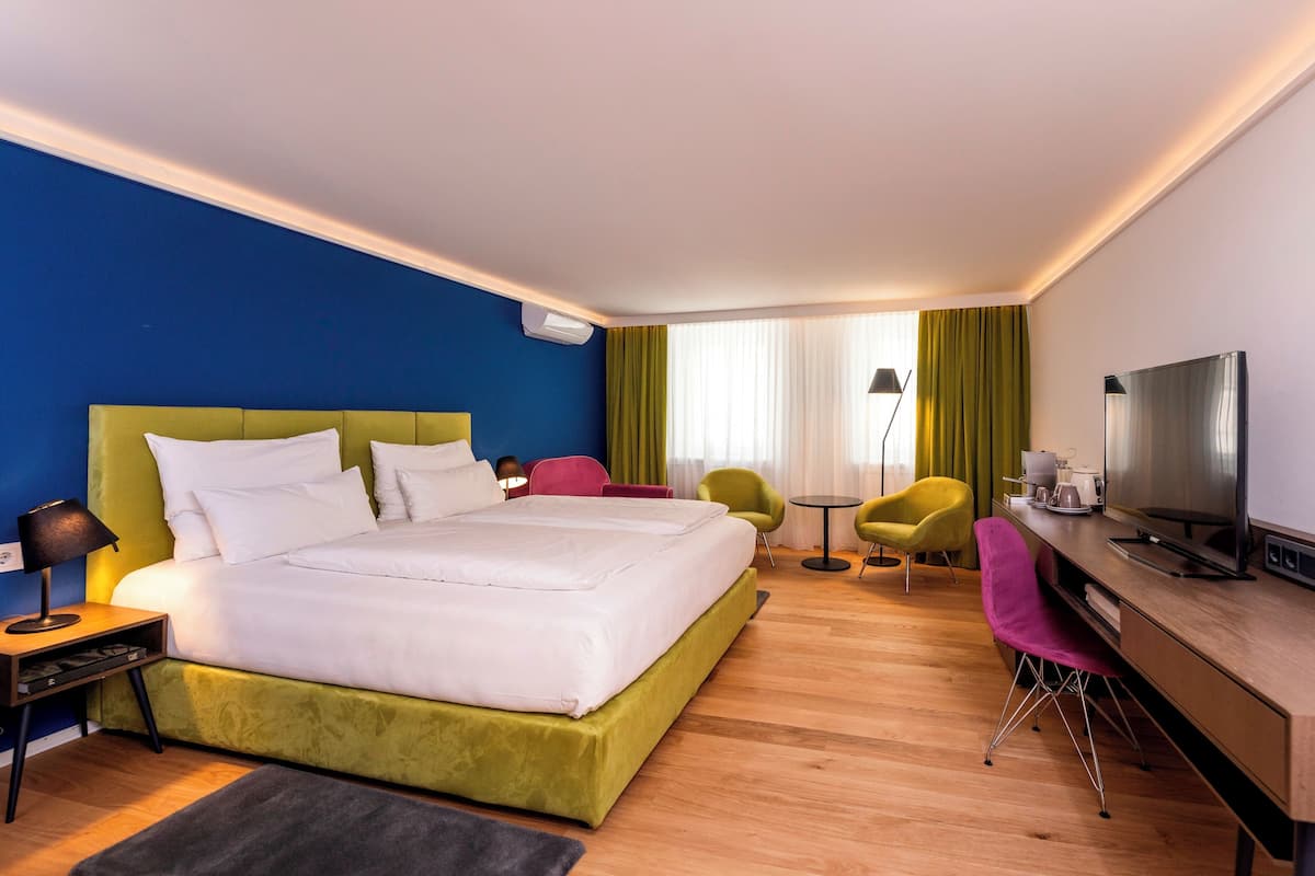 A hotel room with a large bed, a desk with a TV, a chair, and a small table with a lamp.
