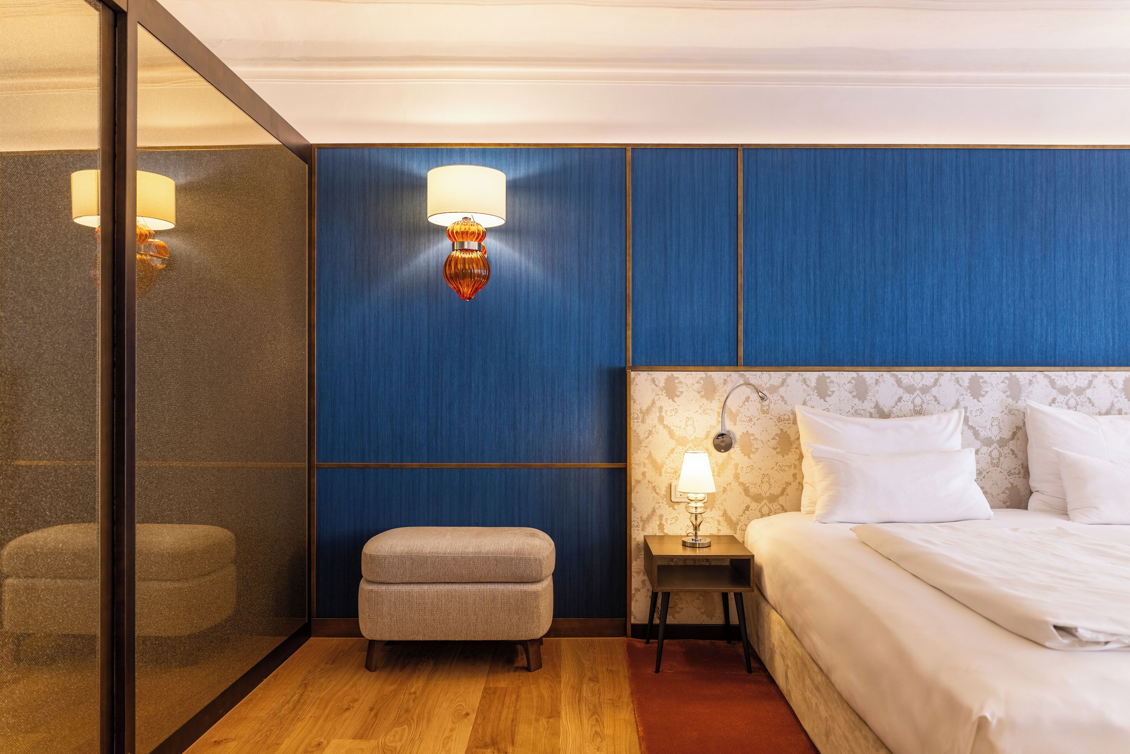 A hotel room with a bed, a nightstand with a lamp, a small ottoman, and a wall with a textured design.