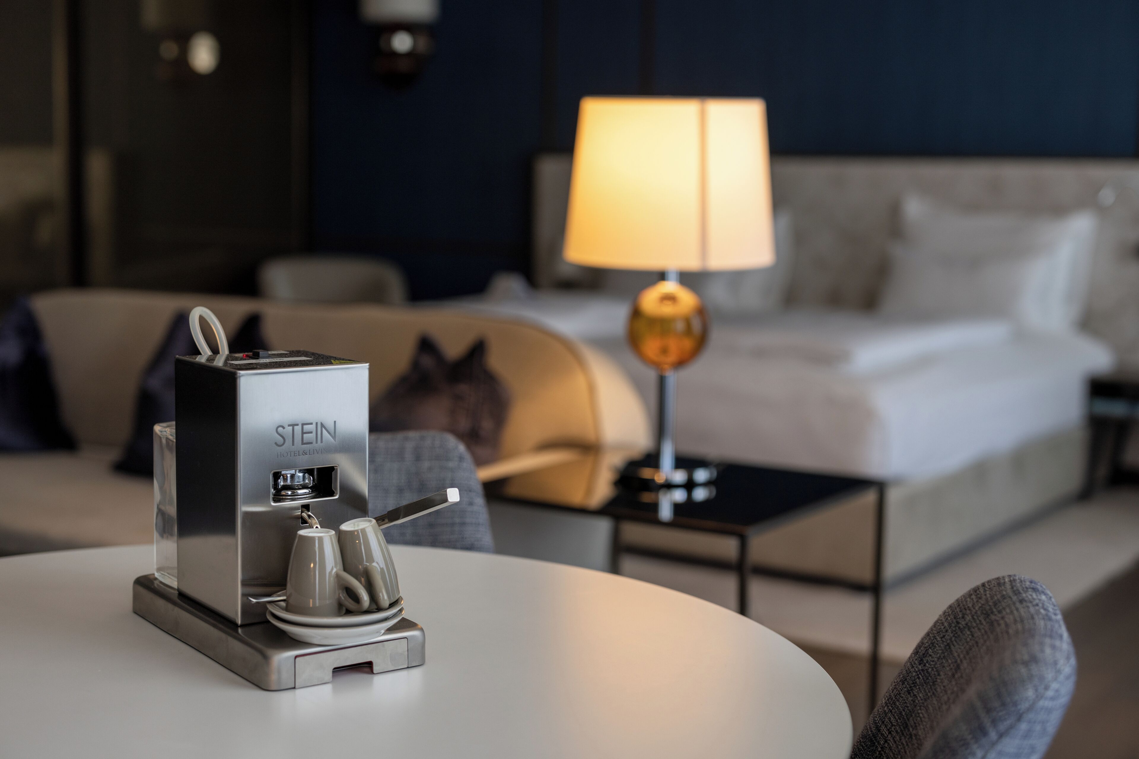 A hotel room with a bed, a lamp, a coffee machine, and a sofa.
