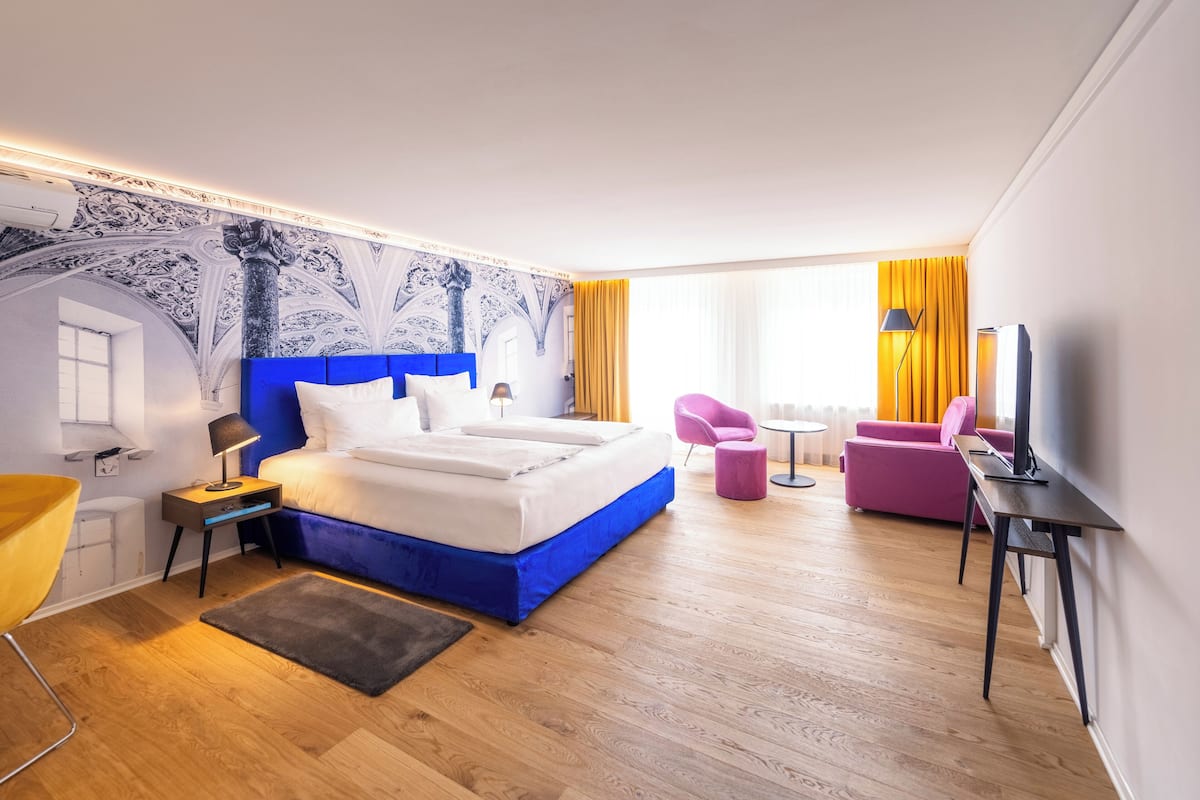 A modern hotel room with a large bed, a desk, a chair, and a TV.