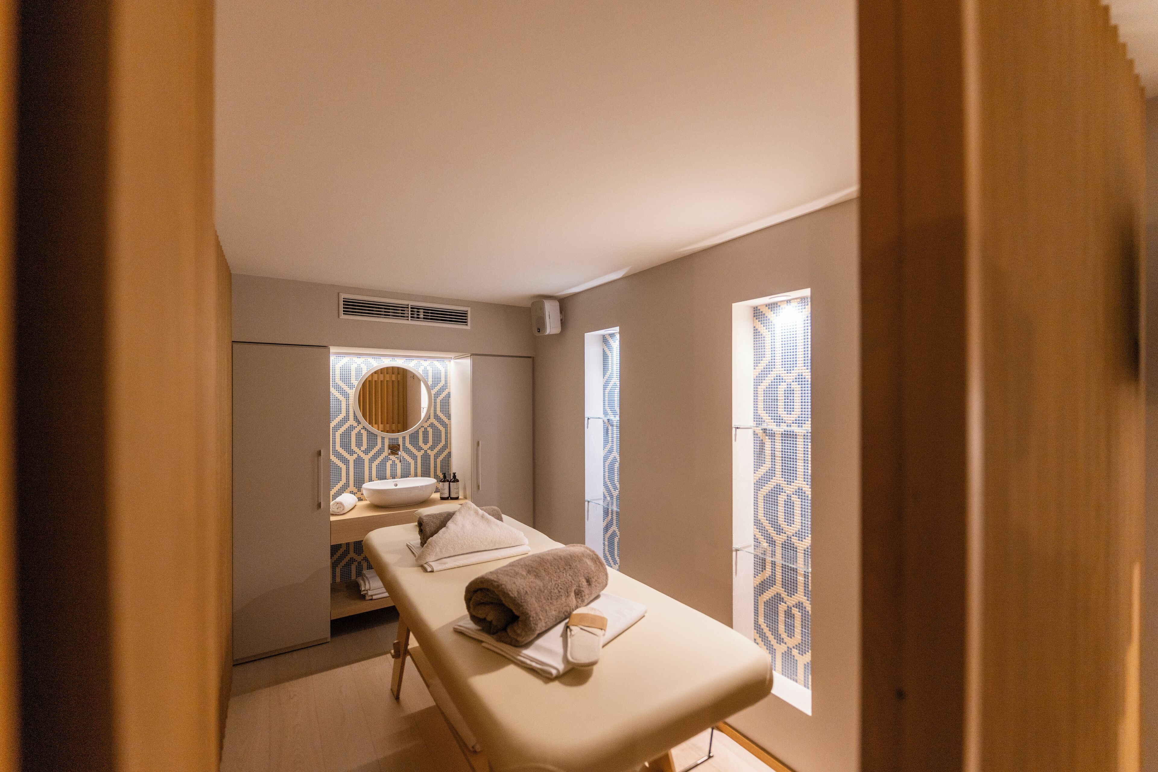 Ein moderner Wellnessraum mit Massageliege, Waschbecken und fensterartigen Verzierungen.