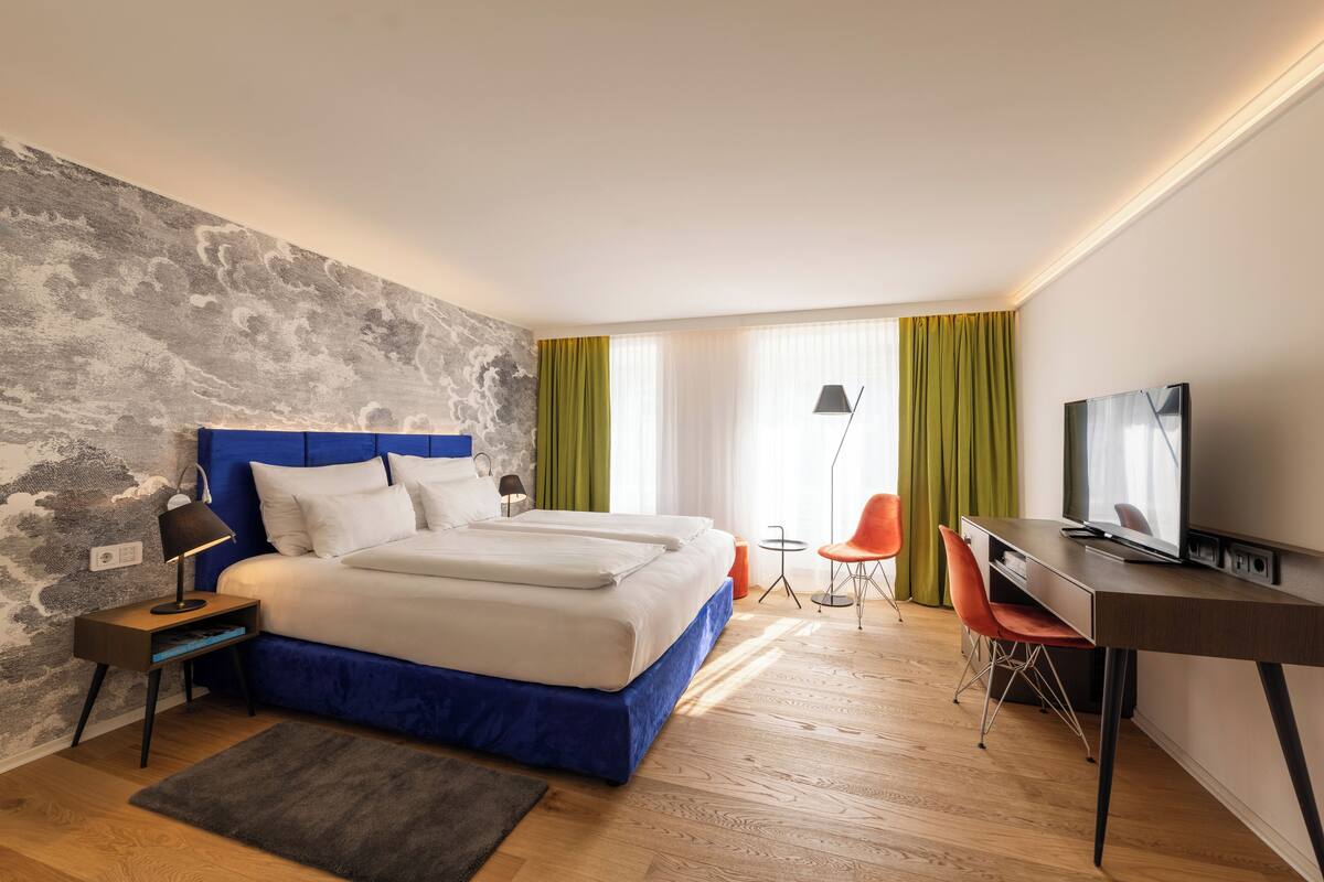 A modern hotel room with a large bed, a desk, a television, and a small table with chairs.
