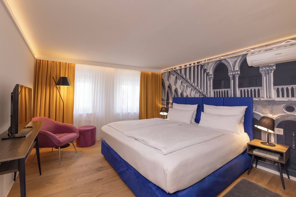 A hotel room with a large bed, a desk, a chair, and a wall mural.