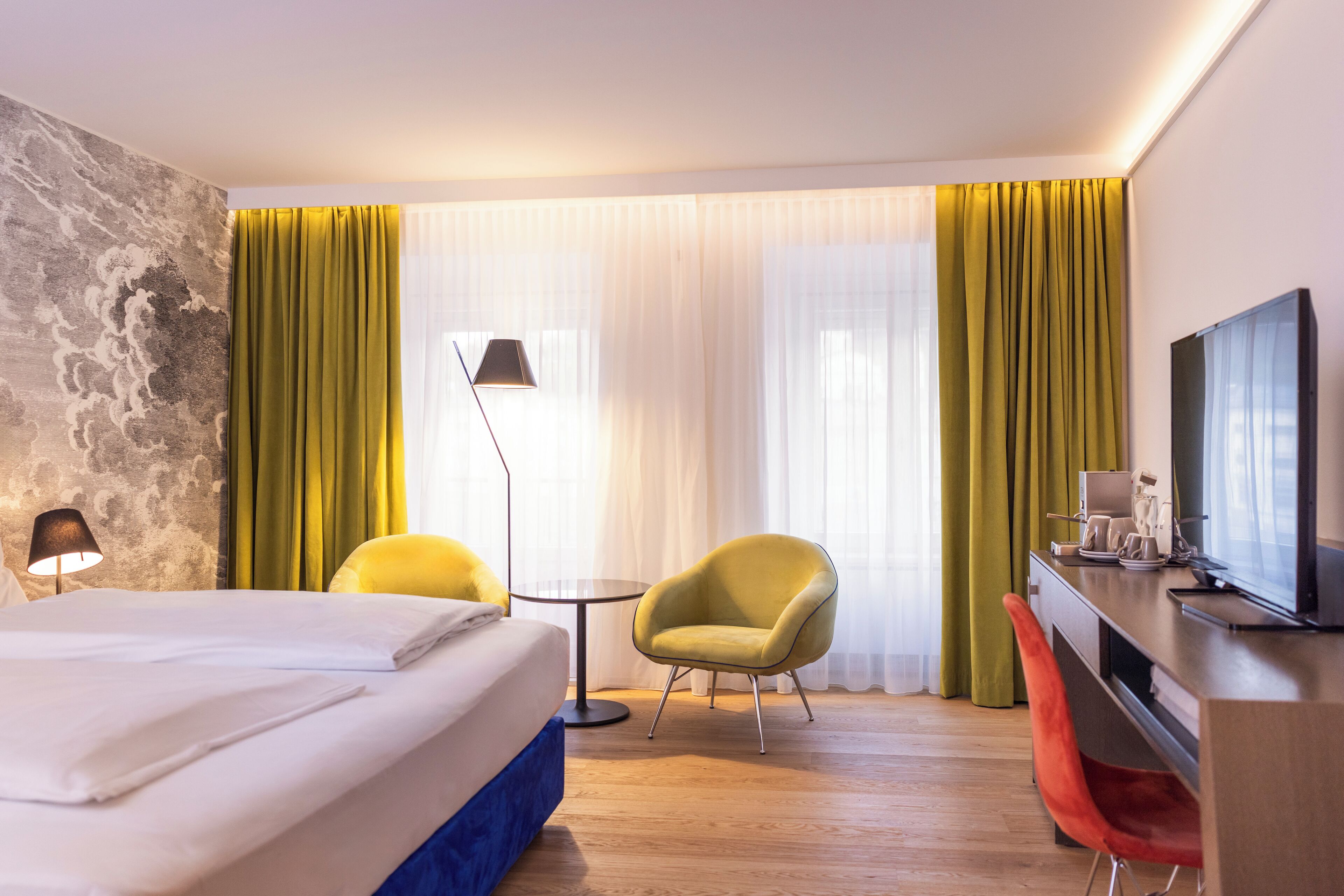 A hotel room with a bed, a desk with a TV, a chair, a lamp, and a window with curtains.