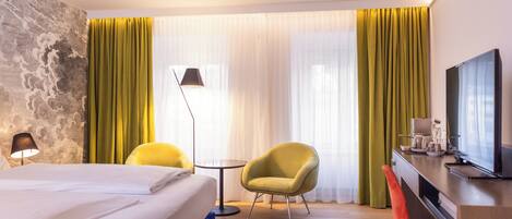 A hotel room with a bed, a desk with a TV, a chair, a lamp, and a window with curtains.