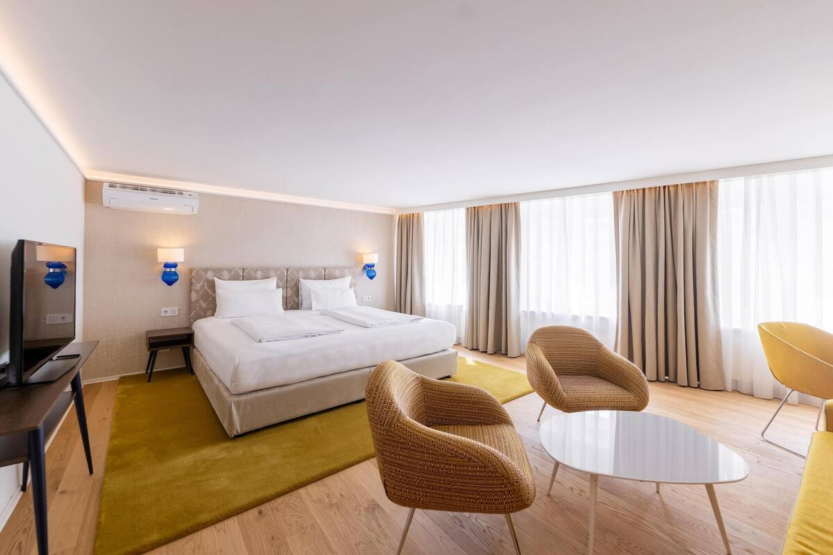 A hotel room with a bed, two chairs, a small table, a TV, and curtains.