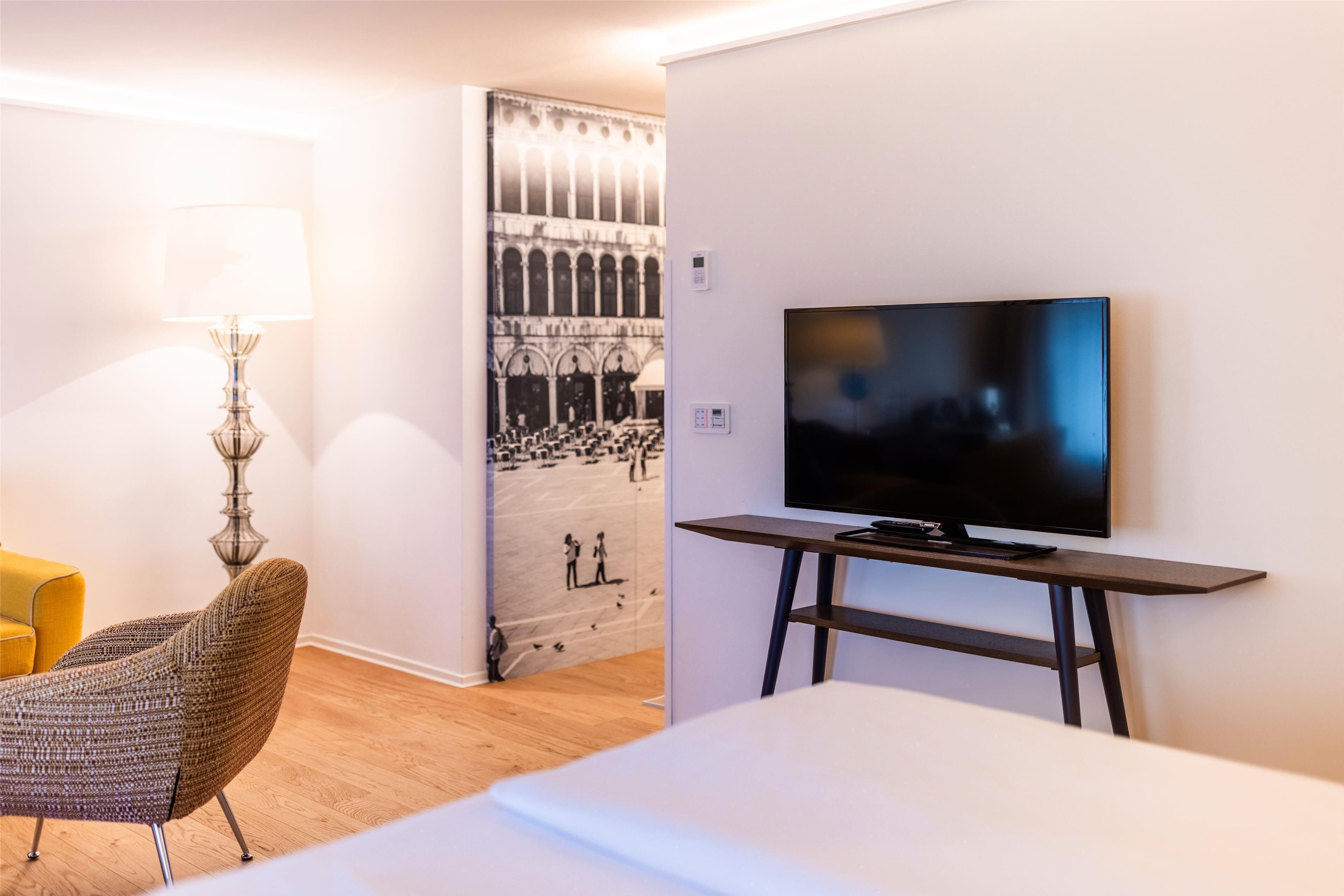 A hotel room with a television on a stand, a lamp, a chair, and a wall with a mural.