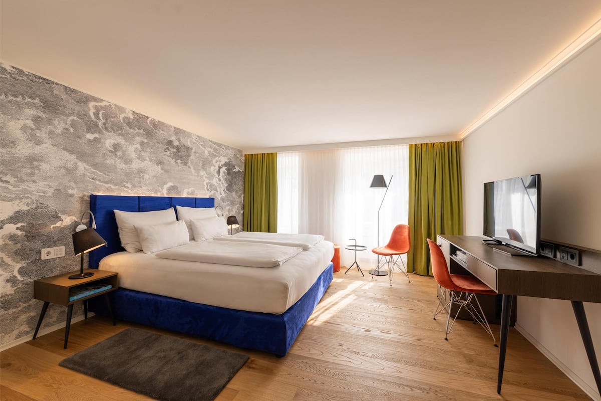 A modern hotel room with a large bed, a desk, a TV, and a small table with chairs.