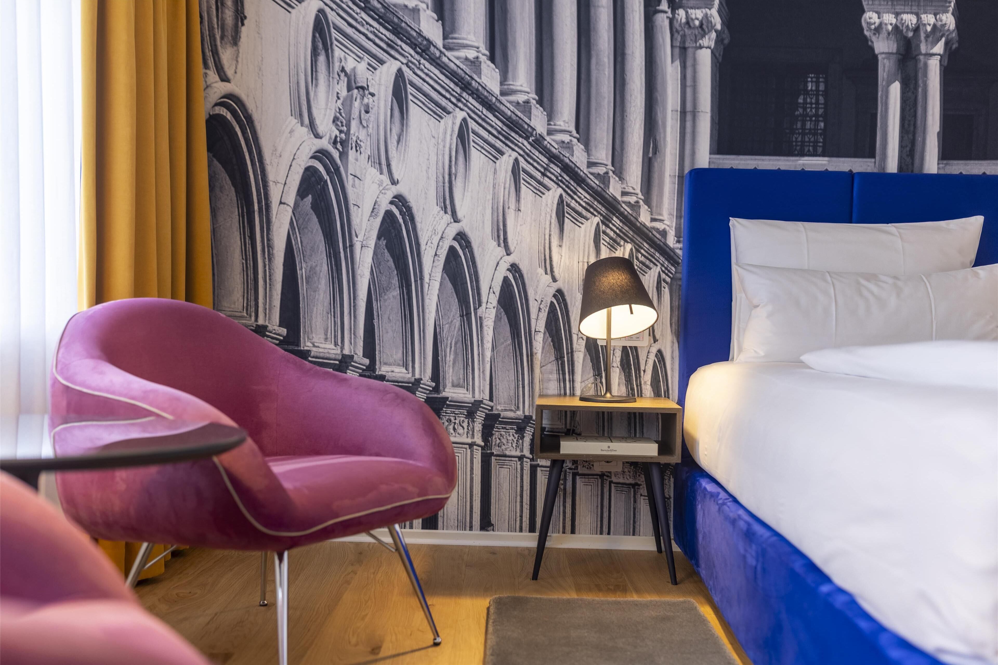 A hotel room with a bed, a desk with a lamp, a purple chair, and a mural of architectural details on the wall.