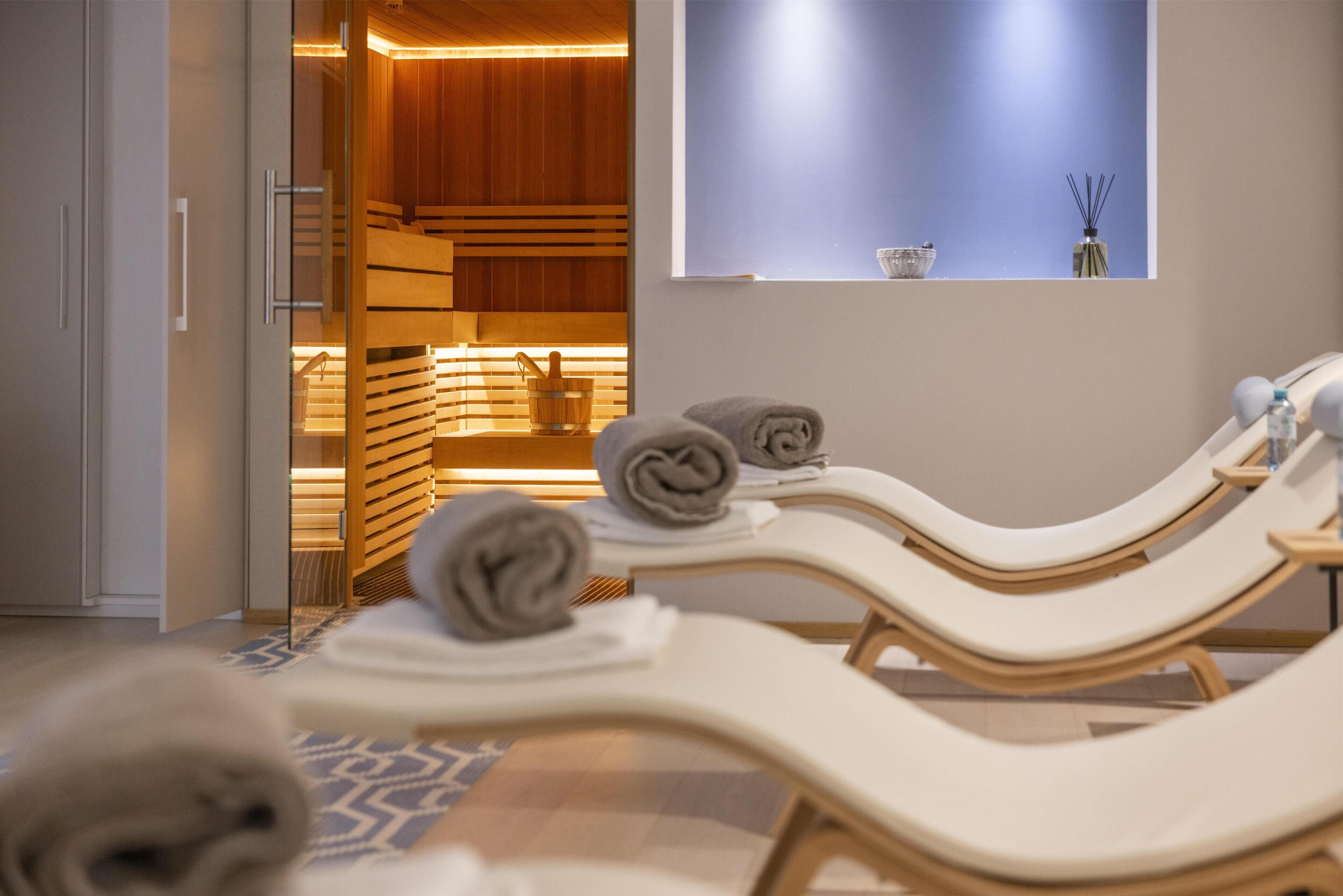 Ein Wellnessbereich mit Holzliegen, Handtüchern und einer Sauna aus Holz, die durch einen offenen Türrahmen sichtbar ist.