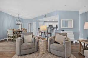 A living room with two grey armchairs, a coffee table, a dining area with a wooden table and chairs, and a kitchen with white cabinets and appliances.