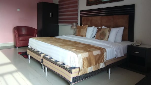 A bedroom with a large bed, a red chair, a wardrobe, and a painting on the wall. - Jesuit Continental Suites (Abuja)