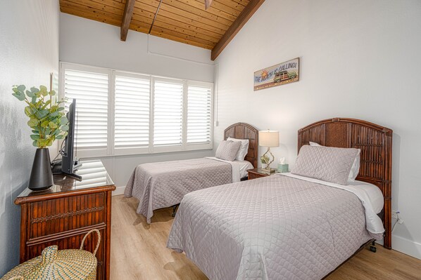 Une chambre avec deux lits, une tête de lit en bois, une table de chevet avec une lampe, une télévision et une plante.