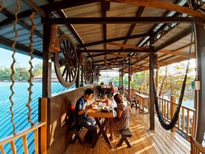 A wooden deck with tables and chairs, large wheels on the ceiling, and a view of the water. - Dream House Guest House & Restaurant (Ngwe Saung)