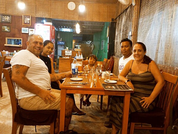 A group of people seated at a wooden table in a restaurant with a rustic interior, including exposed brick walls and wooden furniture. - Dream House Guest House & Restaurant (Ngwe Saung)