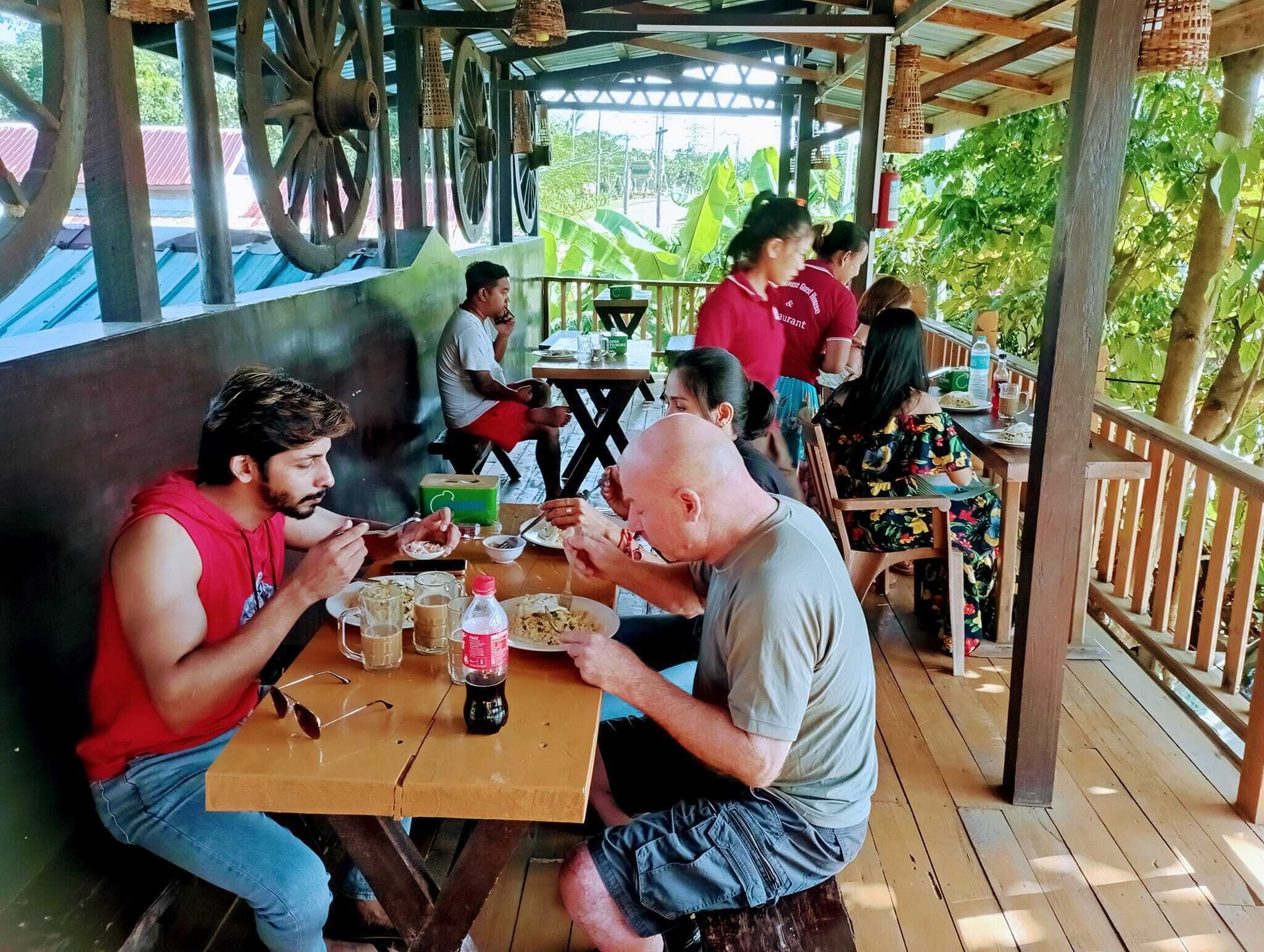 A group of people dining at an outdoor wooden table with a rustic design, surrounded by greenery and a covered area with metal beams and wheels.