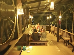Un restaurante con una terraza de madera larga, mesas y luces colgantes.