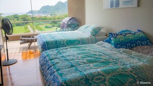 Camera da letto con due letti, un'ampia finestra vista su vegetazione e colline, una ventola a piantana e un piccolo tavolino.