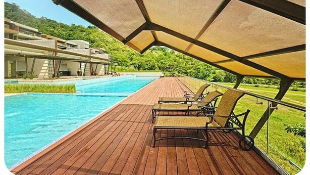 Un solarium in legno con poltrone relax che si affaccia su una piscina, immerso nel verde con un edificio sullo sfondo.