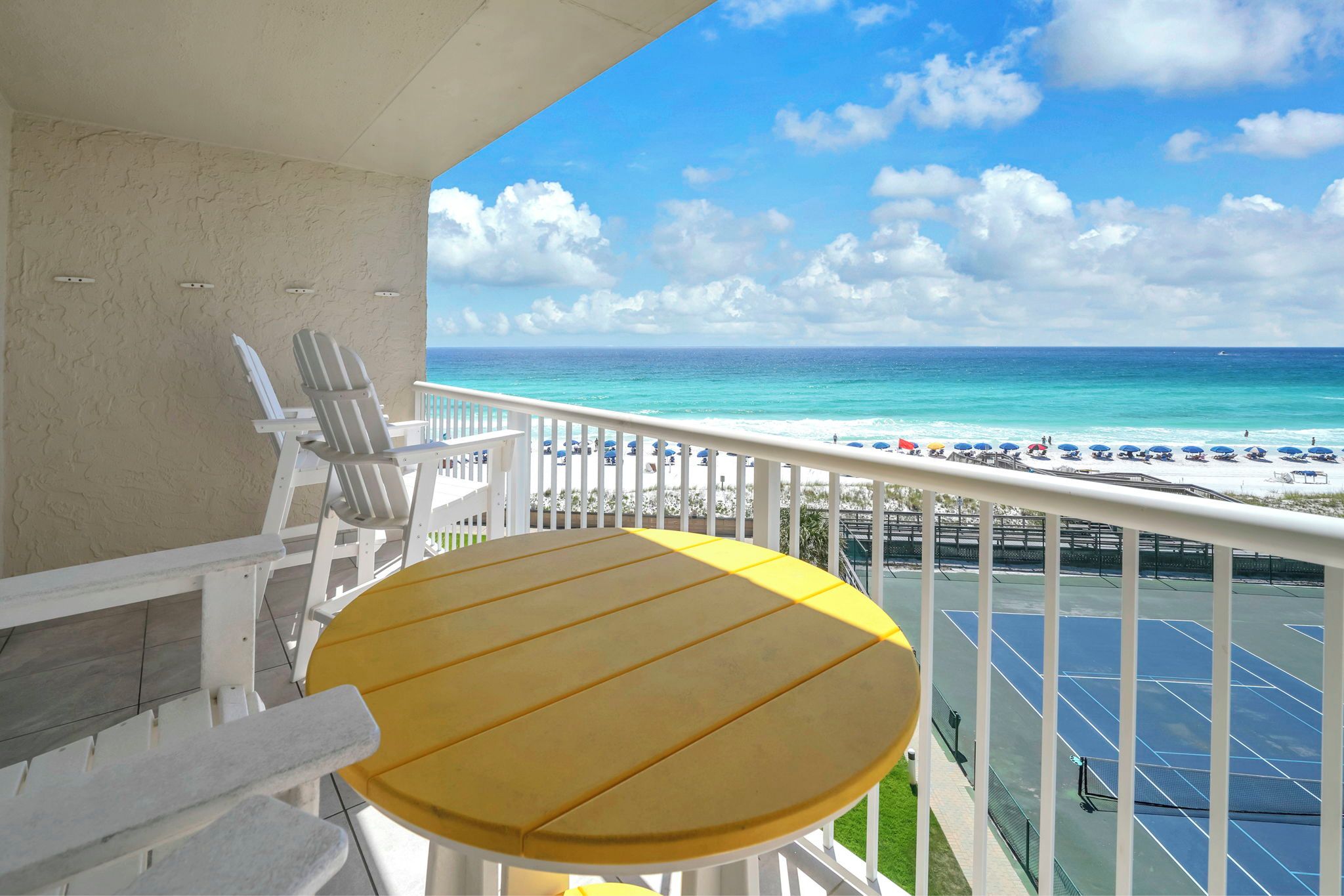 Un balcon avec une table en bois et des chaises blanches, donnant sur une plage parsemée de parasols et un court de tennis.