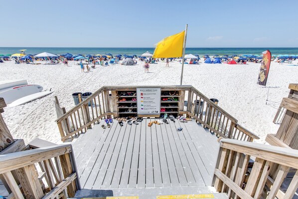 A wooden deck with a sign indicating beach warning flags, a beach with umbrellas, and a clear view of the ocean.