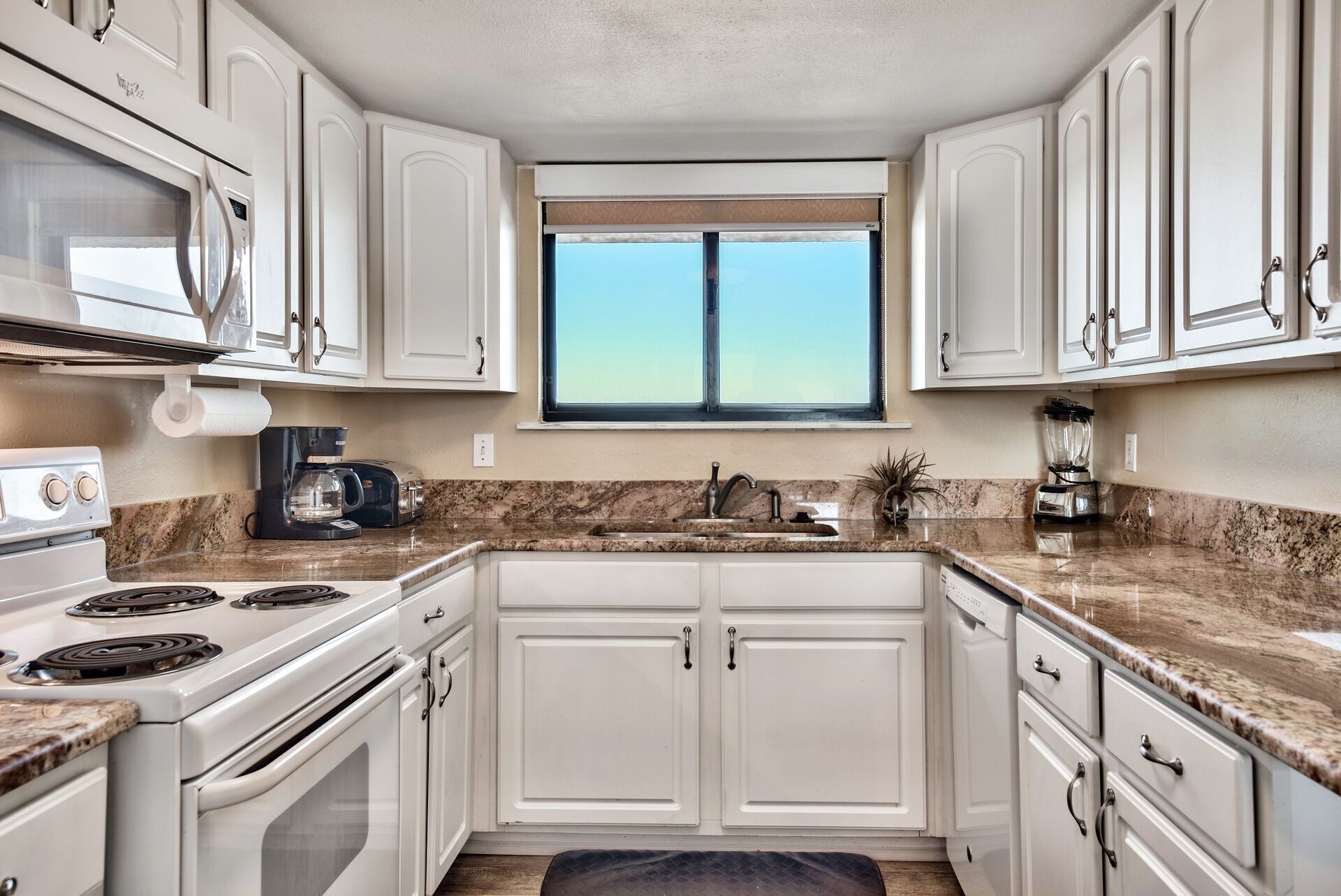 A kitchen with white cabinets, a stove, a microwave, a window, and a sink.
