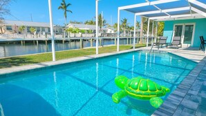 Ein klares, blaues Schwimmbecken mit einem grünen, aufblasbaren Schildkröten-Schwimmpfädchen, umgeben von einer überdachten Liegefläche mit Liegestühlen und Blick auf eine Marina.