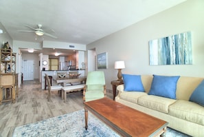 A living room with a wooden coffee table, beige sofa with blue cushions, a dining area with chairs, and a kitchen in the background. - 1703 - 3 Bedroom 3 Bath. Master Bedroom on the Gulf (Panama City Beach)