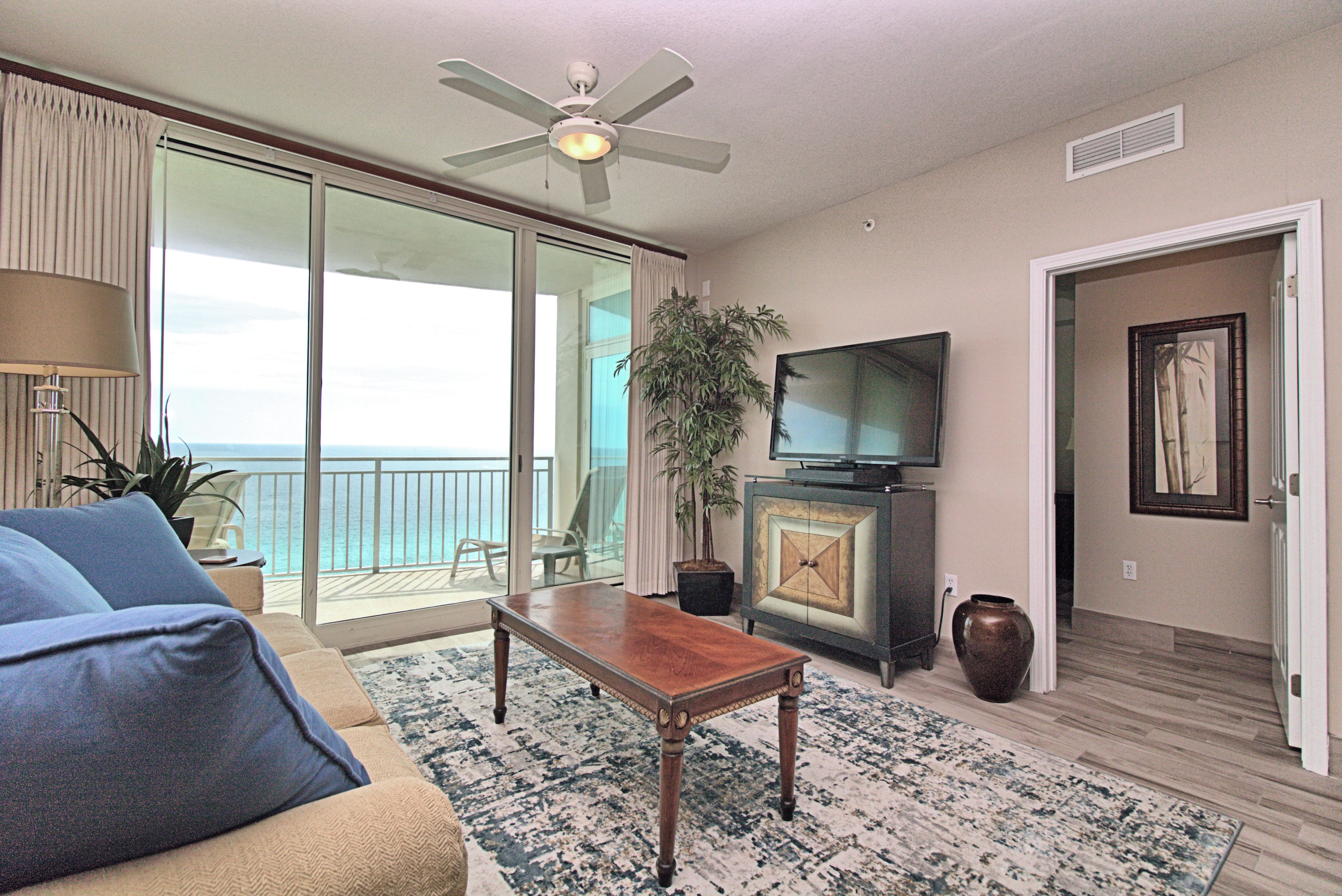 A living room with a sofa, a coffee table, a television, a plant, and a view of the ocean through a sliding glass door.