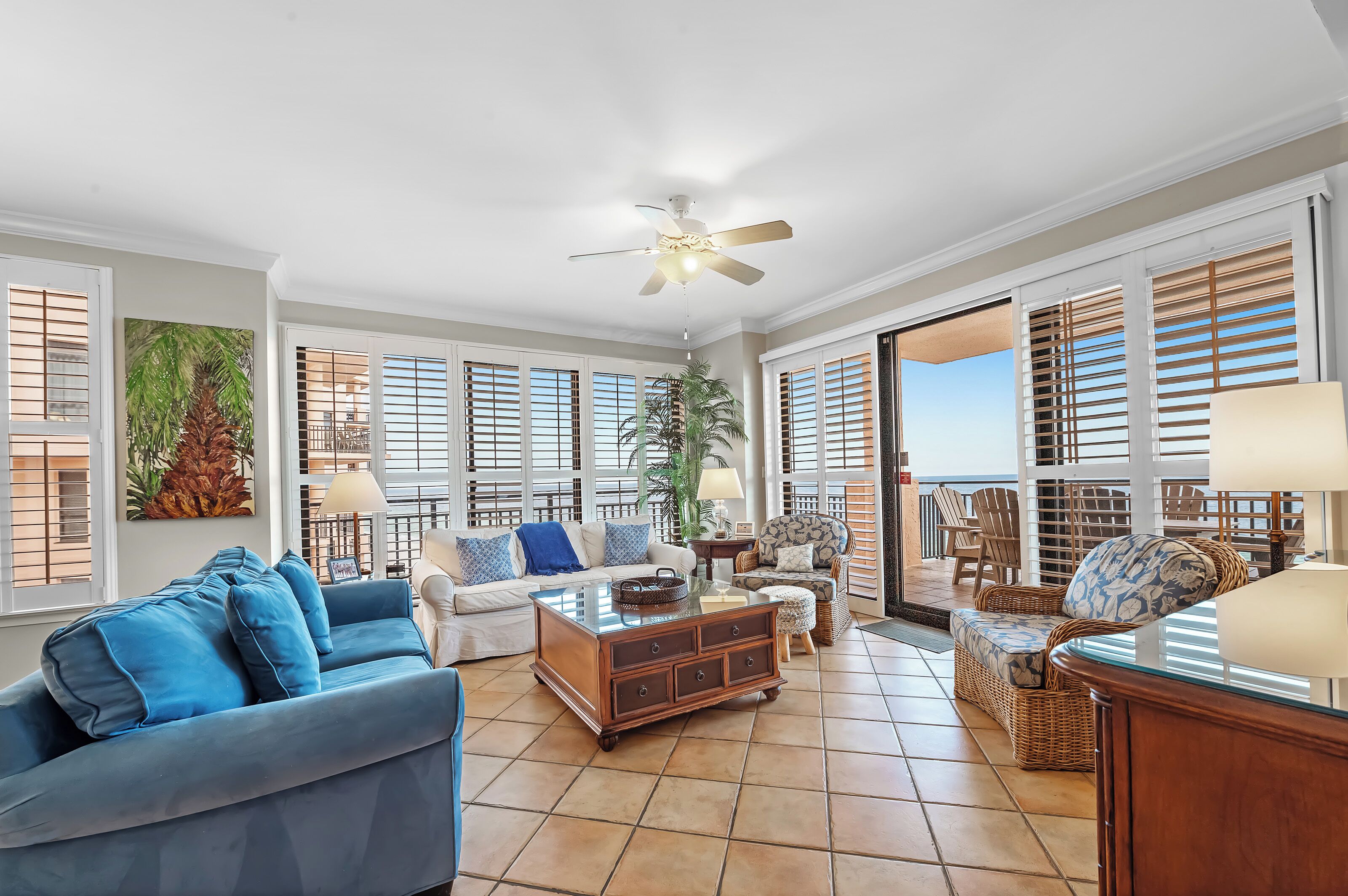 A spacious living room with a blue sofa, a wooden coffee table, wicker chairs, and large windows with shutters.