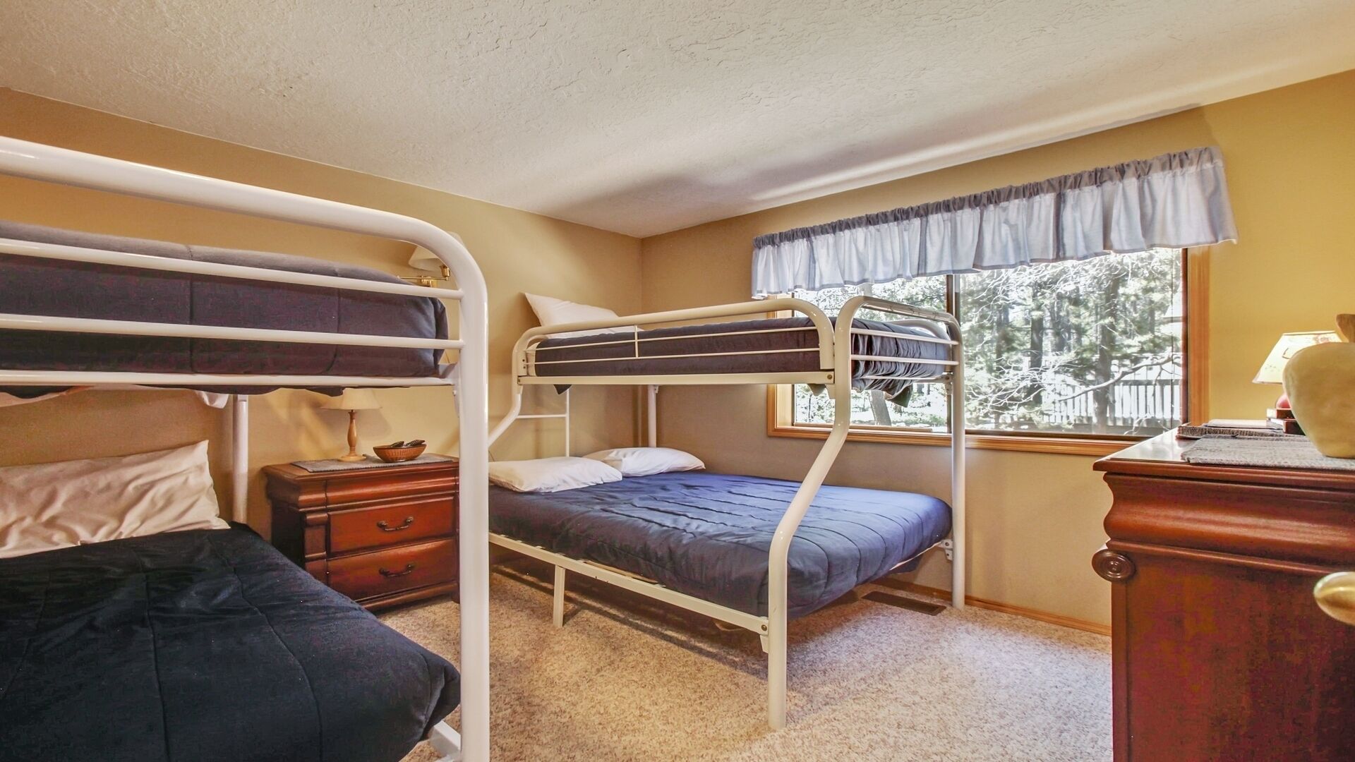 A dormitory room with bunk beds, a small wooden dresser, a nightstand, and a window with a view of trees.