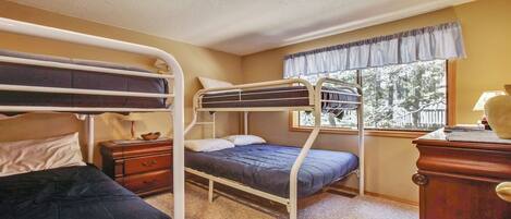 A dormitory room with bunk beds, a small wooden dresser, a nightstand, and a window with a view of trees.