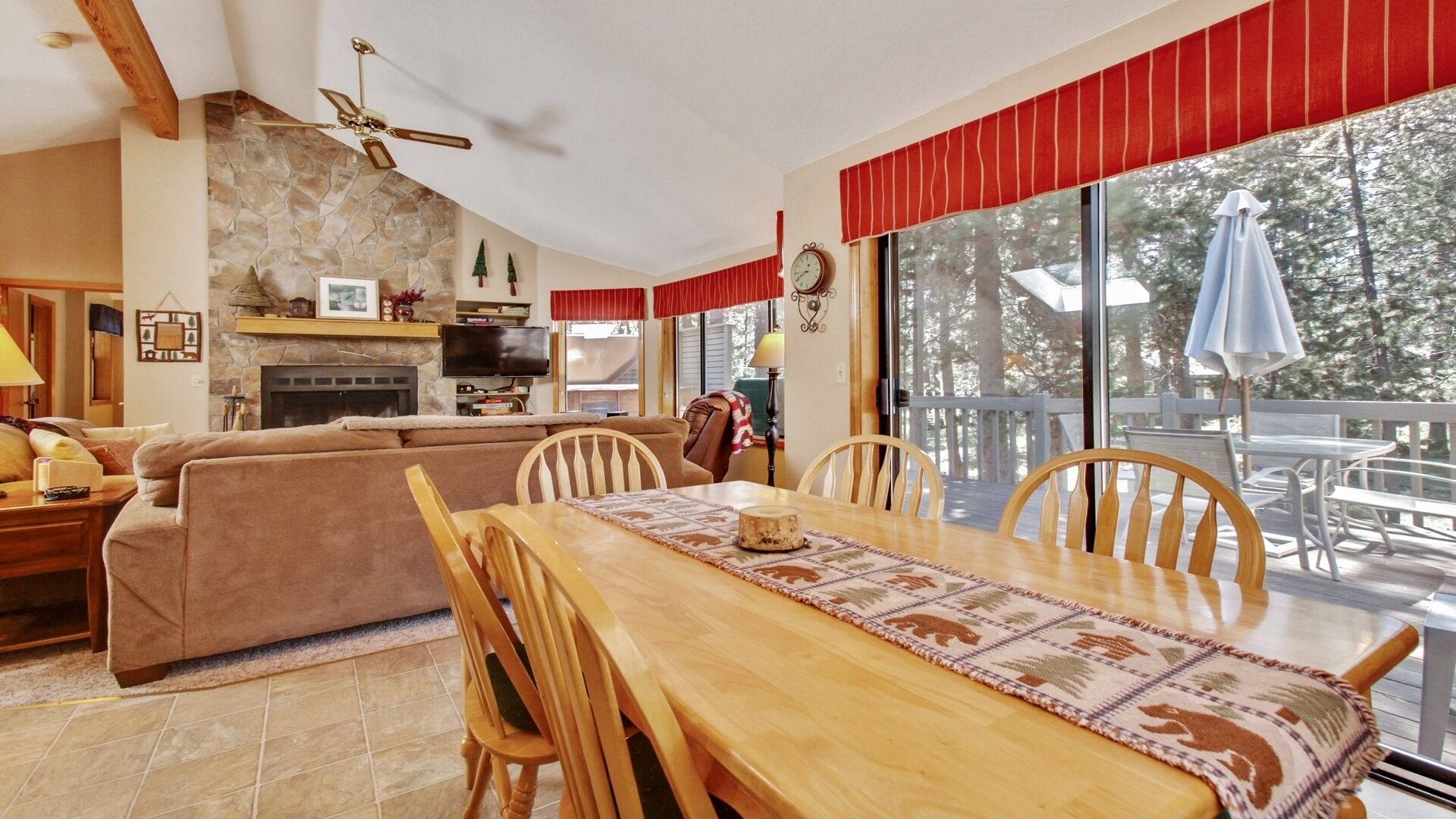 Un salon chaleureux avec une cheminée, un coin repas avec une table et des chaises, et une grande fenêtre donnant sur la forêt.
