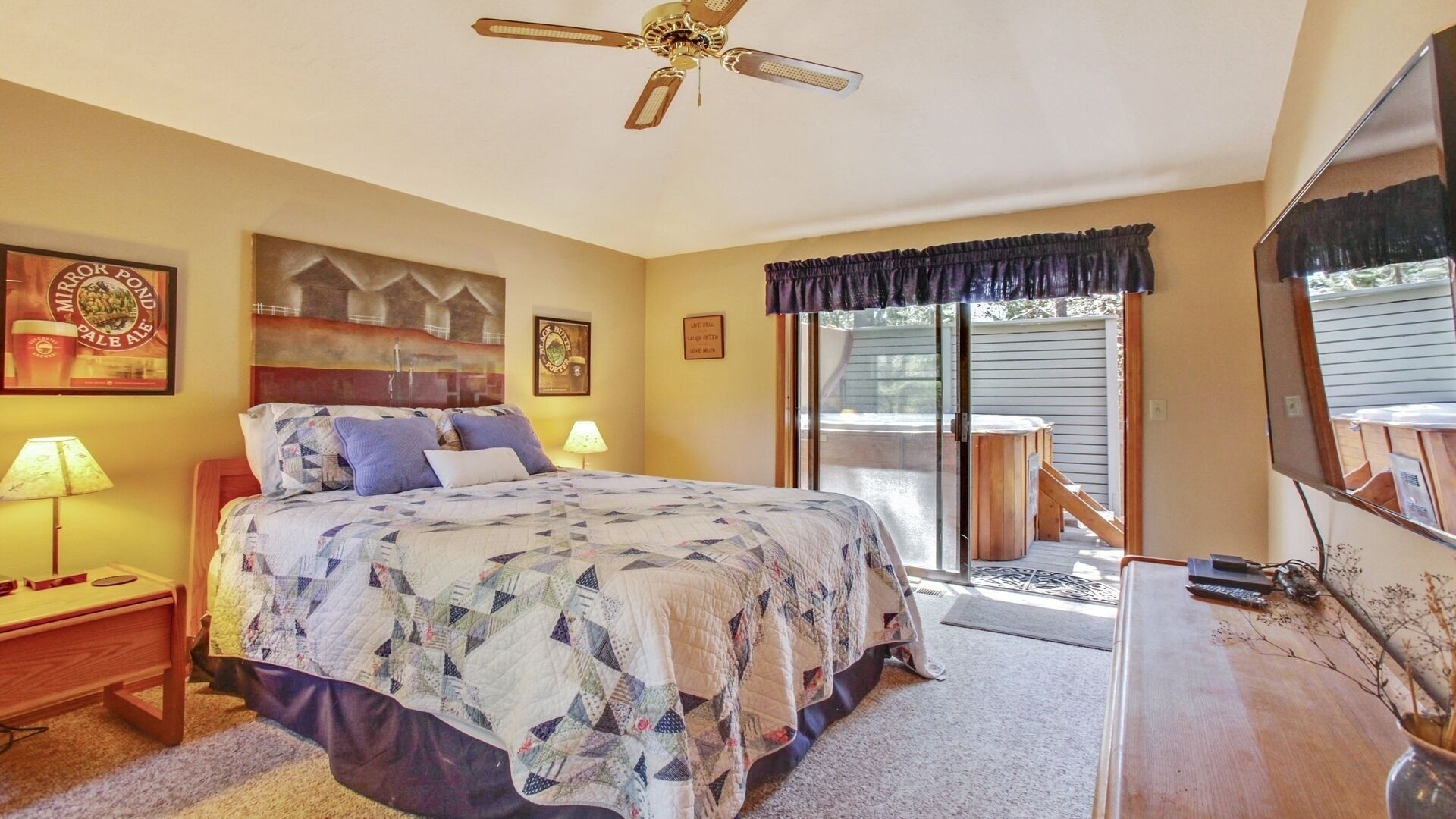 A bedroom with a bed, a nightstand, a lamp, a ceiling fan, a window with a view, and a framed picture on the wall.