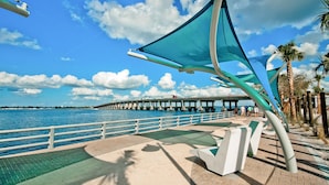A waterfront promenade with a bridge in the background, a railing, and palm trees.
