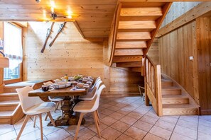 A wooden interior with a dining area, chairs, a table with various items, and a staircase.