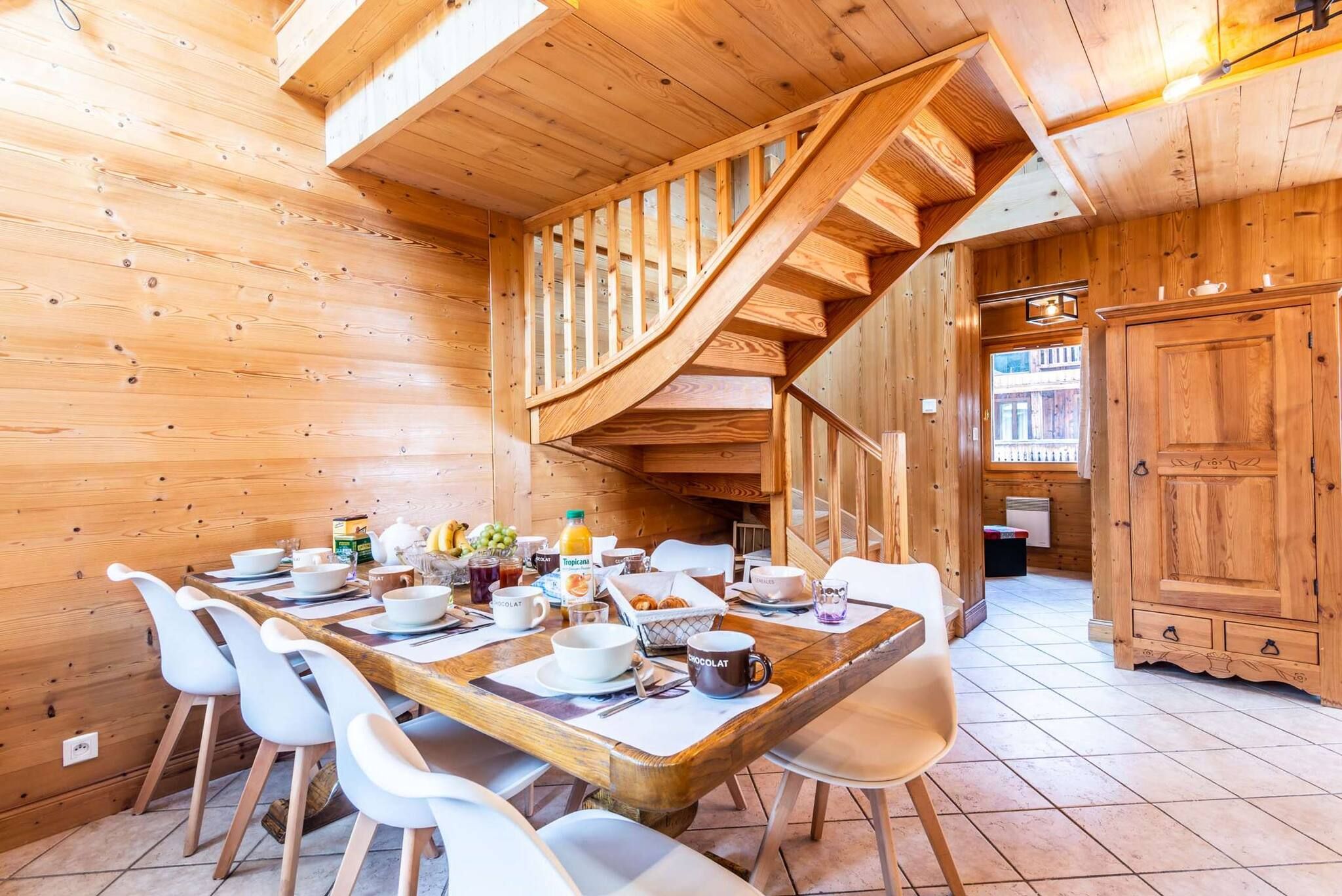 A wooden interior with a dining area featuring a long table, white chairs, and a staircase.