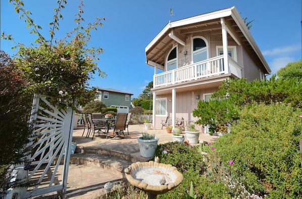 A two-story house with a balcony, surrounded by a garden with potted plants and a bird bath.