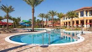 A swimming pool with palm trees, a water slide, and a building with red umbrellas.