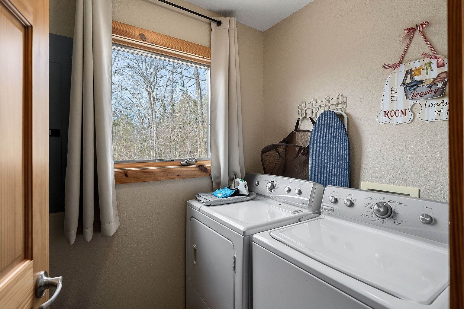 A compact laundry room with two washing machines, a drying rack, and a window with curtains.