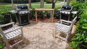 Een terras buiten met twee barbecues, een houten tafel en twee witte stoelen.