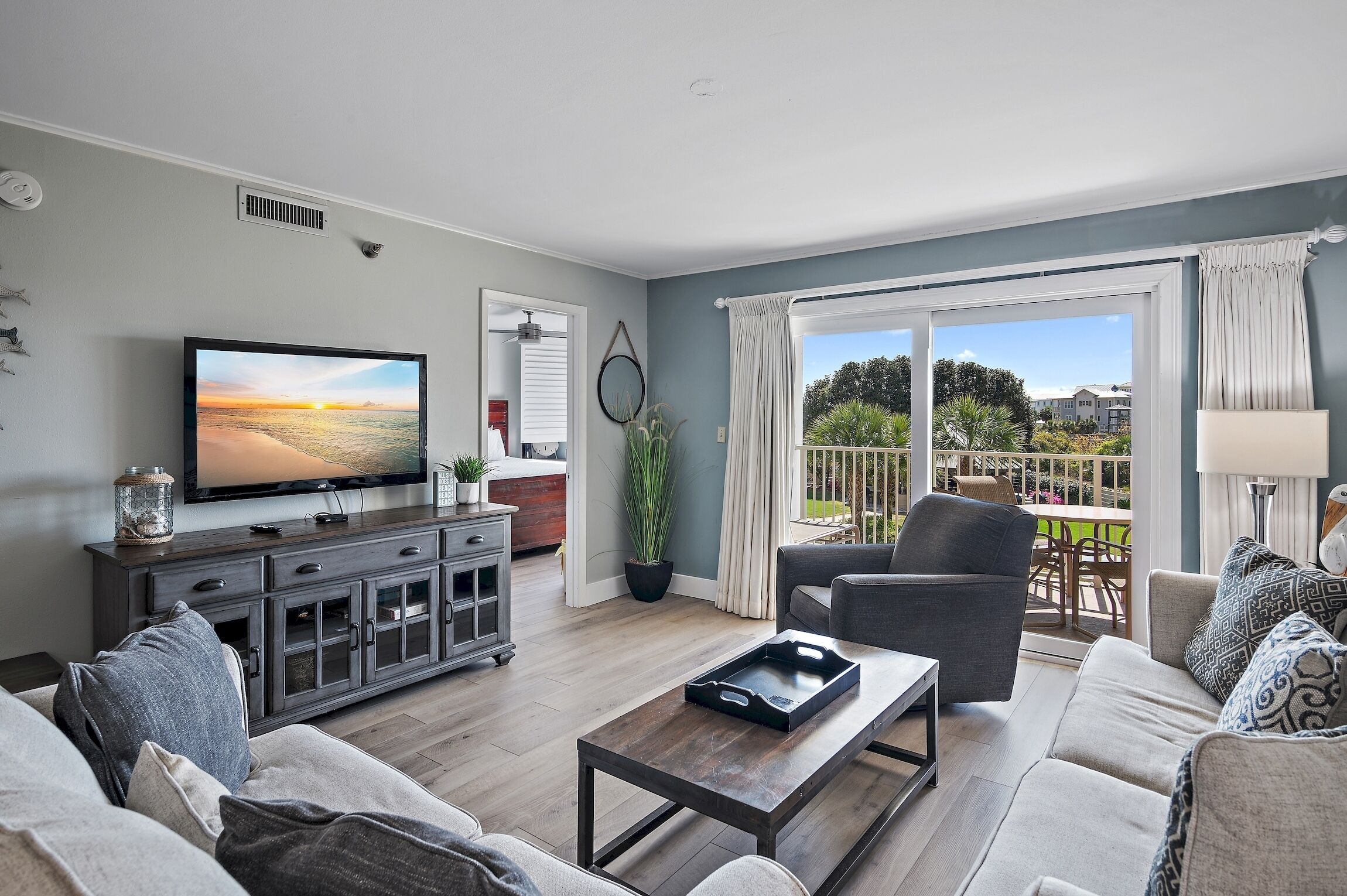 A living room with a flat-screen TV, a dark wooden cabinet, a coffee table, a grey sofa, and a view of a balcony with outdoor furniture.
