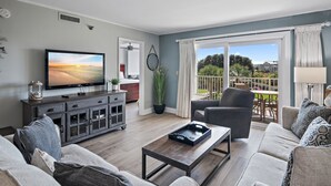 A living room with a flat-screen TV, a dark wooden cabinet, a coffee table, a grey sofa, and a view of a balcony with outdoor furniture. - Great Views, Coastal Style & Fun! Platinum Condo – Islander 217 (Destin)